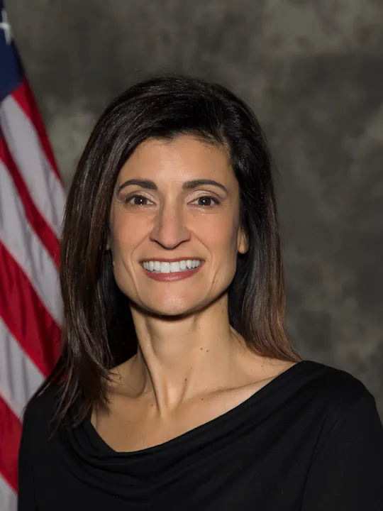 A smiling woman in a black top poses in front of a partially visible American flag.