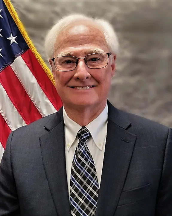 The image features a smiling man in a suit and glasses, standing in front of an American flag.