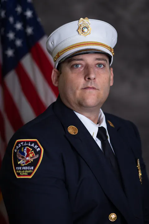 A portrait of a firefighter in uniform with a badge, in front of an American flag.