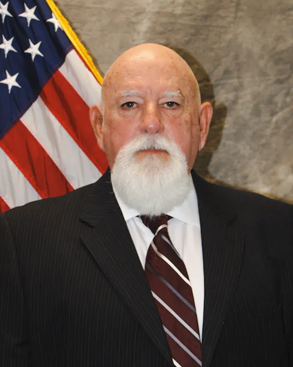 The image features an older man with a prominent white beard, wearing a suit and tie, standing in front of an American flag.