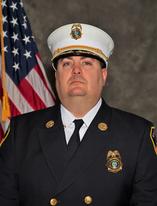 A man in a formal fire department uniform stands in front of an American flag, looking serious and professional.