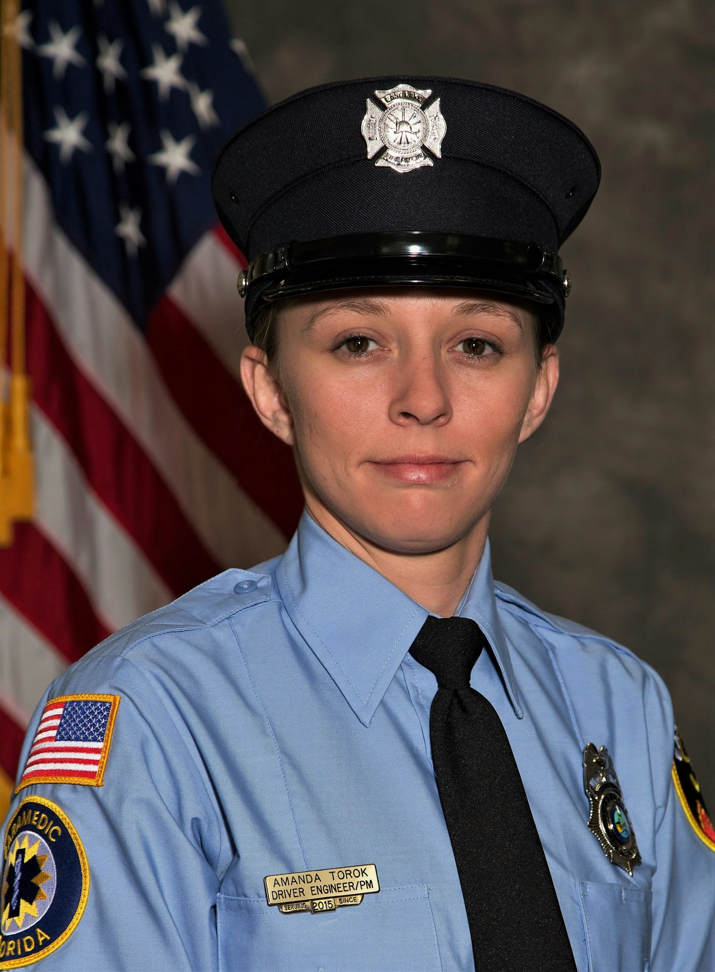 Image of a female firefighter in uniform, with a badge and nameplate, posing in front of an American flag backdrop.