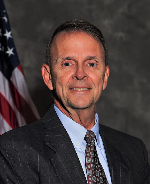 The image shows a middle-aged man in a suit, smiling in front of a gray background with an American flag.