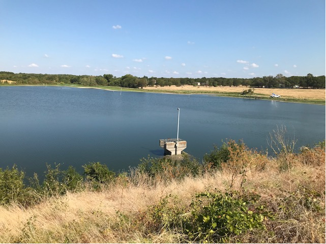 A serene lake view with a small dock, surrounded by grassy terrain and distant trees under a clear blue sky.