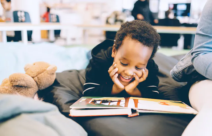 A cheerful child lays on a bed, happily reading a book, with a teddy bear nearby and people in the background.