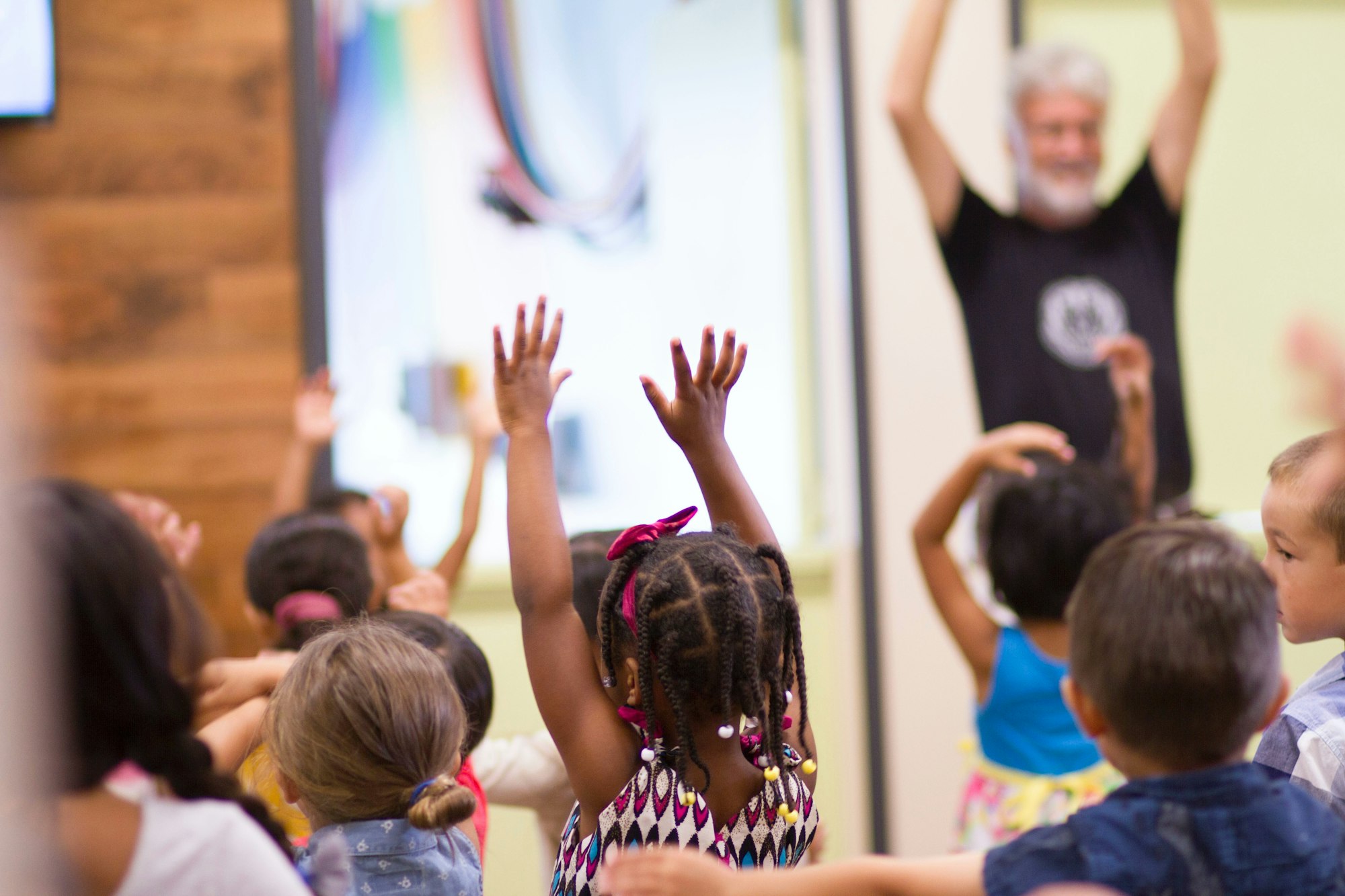 Children are raising their hands in a bright, playful setting while an adult leads in the background.