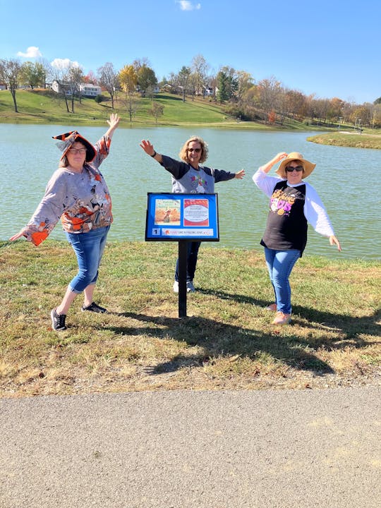 Pictured are 3 Fleming County Library staff surrounding a story board on the story walk trail. There is a lake in the background, along with a clear, blue sky.