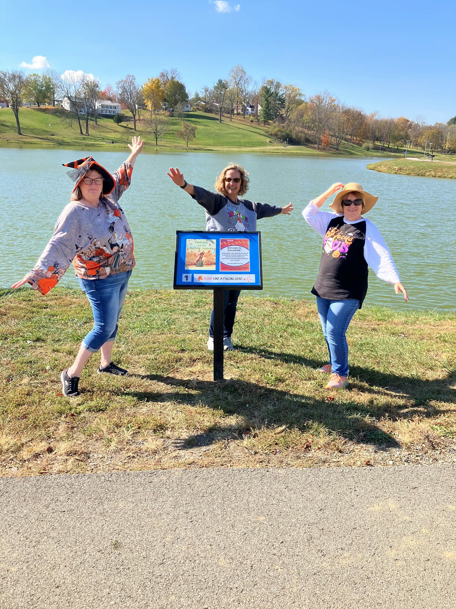 Pictured are 3 Fleming County Library staff surrounding a story board on the story walk trail. There is a lake in the background, along with a clear, blue sky.