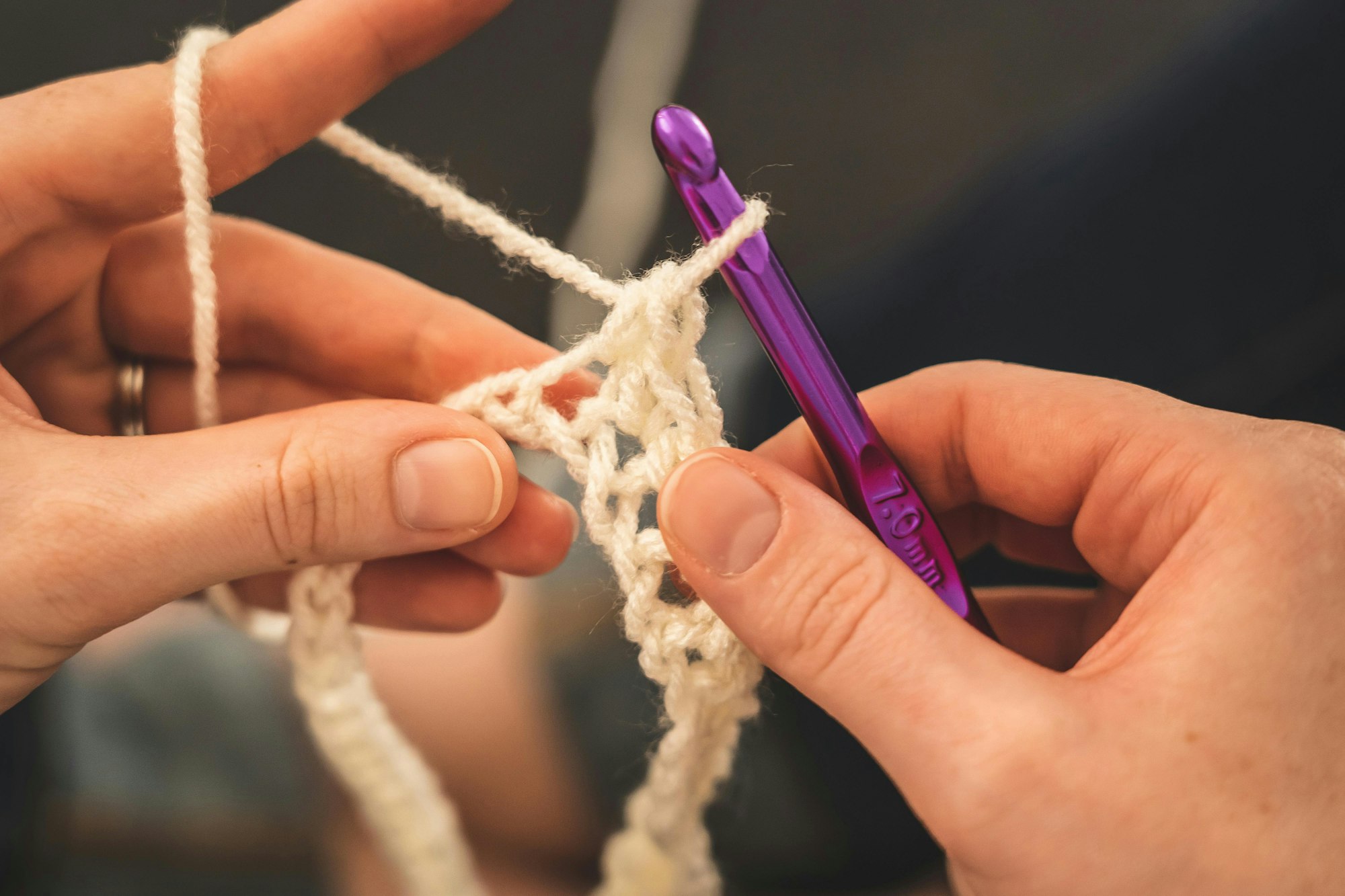 Hands crocheting with white yarn and a purple hook.