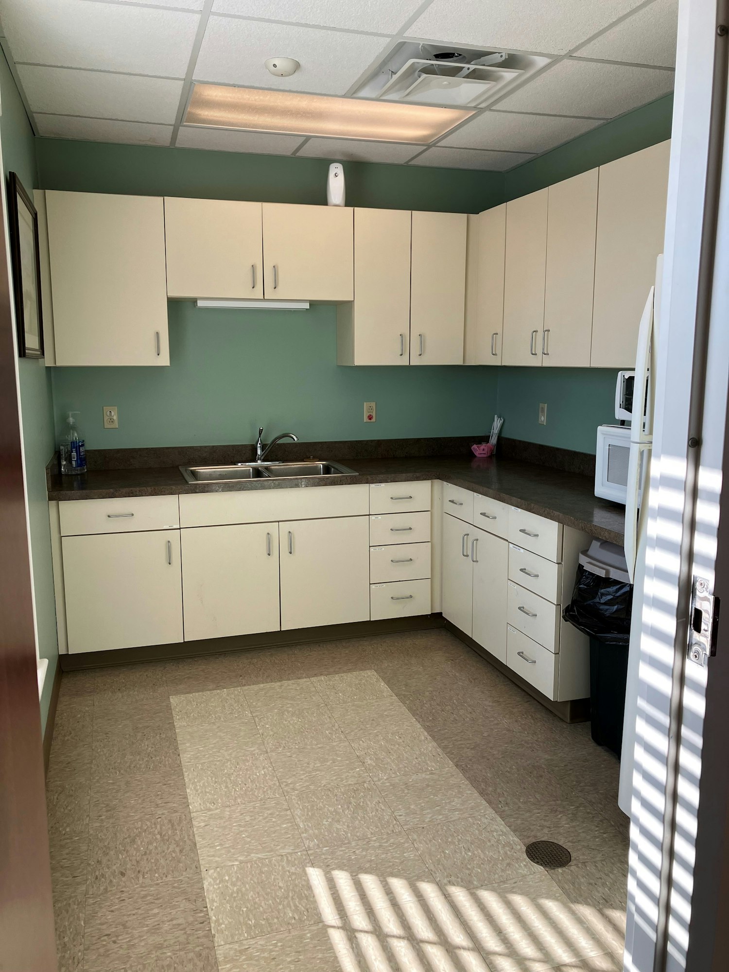 Community Room kitchen with white cabinets, sink, refrigerator and microwave, and one large window casting a shadow on the floor.