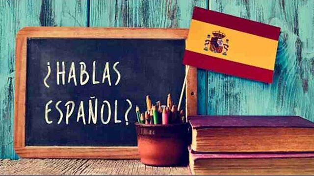 A chalkboard with "¿Hablas español?", a cup of colored pencils, the Spanish flag, and books on a wooden background.