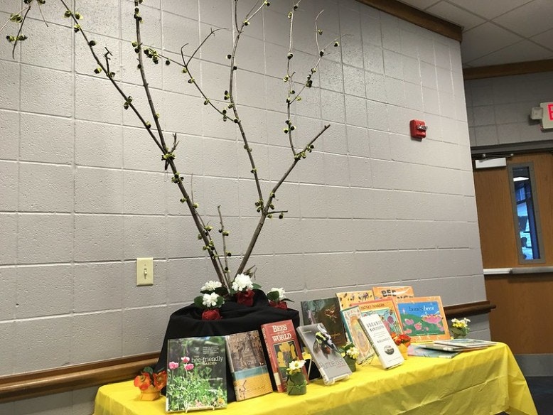 A table with books, a decorative branch arrangement, and flowers against a wall.