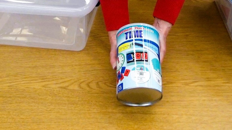 A person holding a can labeled "Math Flash Cards: Time" on a wooden table.