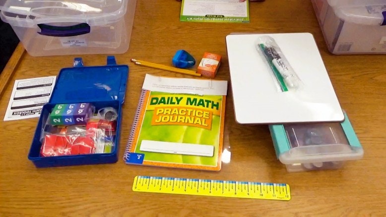 Math practice kit: math journal, ruler, pencil, dice, tape, markers, numbered cubes, and a clear box on a table.