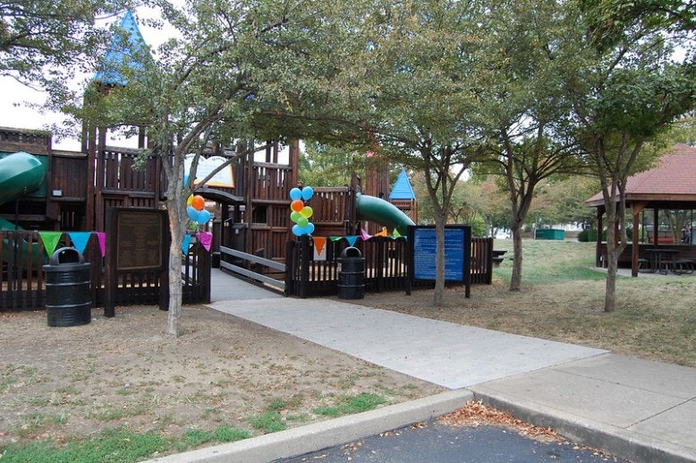 Playground with wooden structures, decorated with colorful balloons and banners. Trees and a pavilion are nearby.