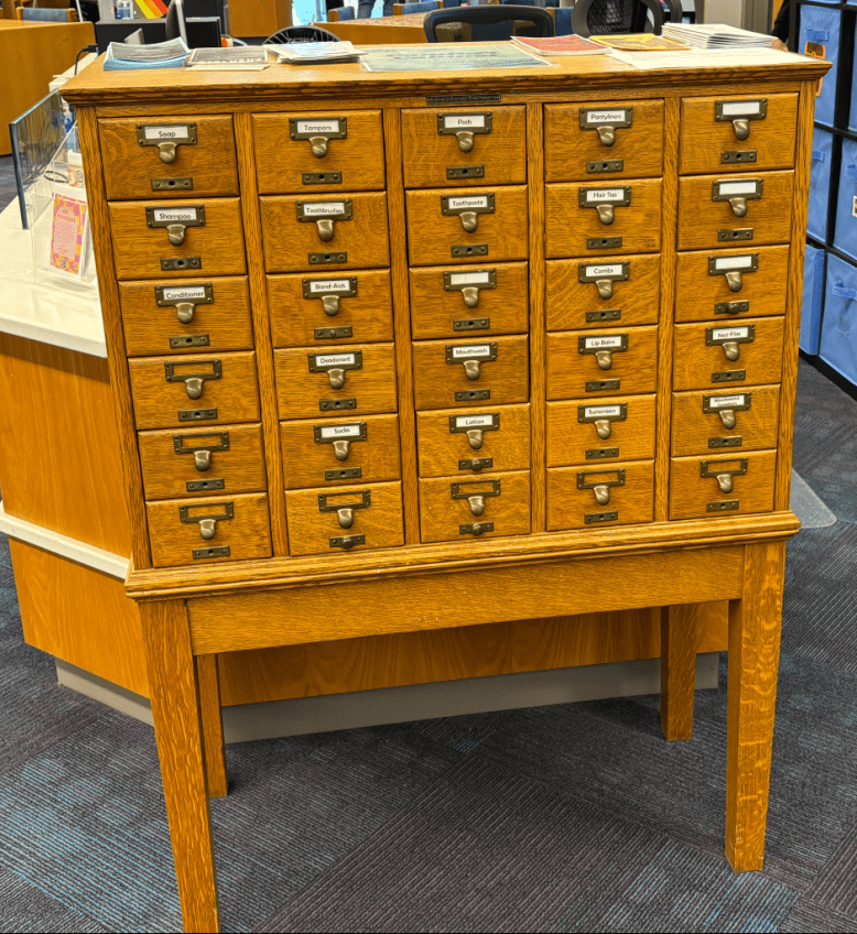 A vintage wooden card catalog with labeled drawers, each listing various personal care items like soap, toothpaste, and deodorant.