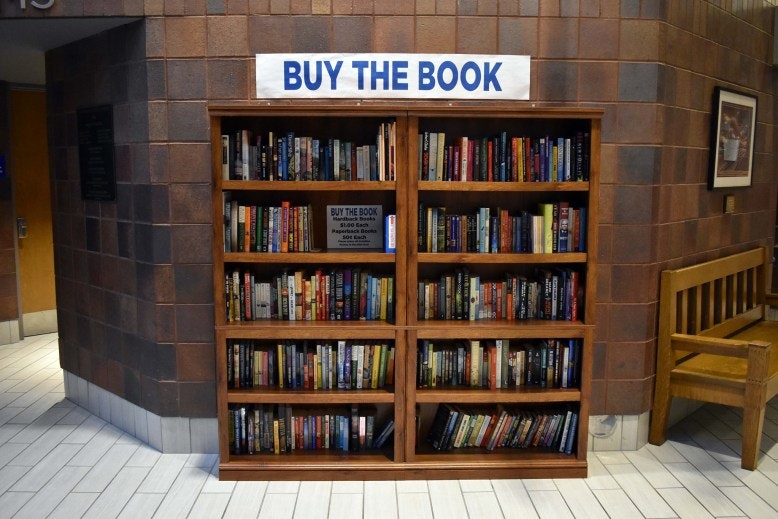 A bookshelf with a sign reading "Buy the Book" filled with various books for sale in a hallway.