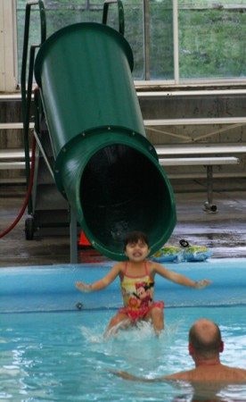 A child in a swimsuit is joyfully sliding down a green waterslide into a pool, with an adult nearby.