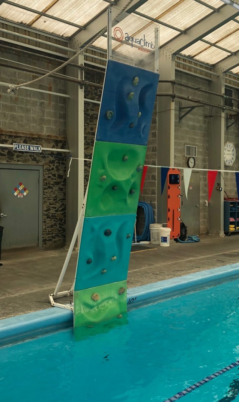 An aqua climbing wall is set up beside a swimming pool, with colorful holds on a slanted surface and swimming lanes visible.