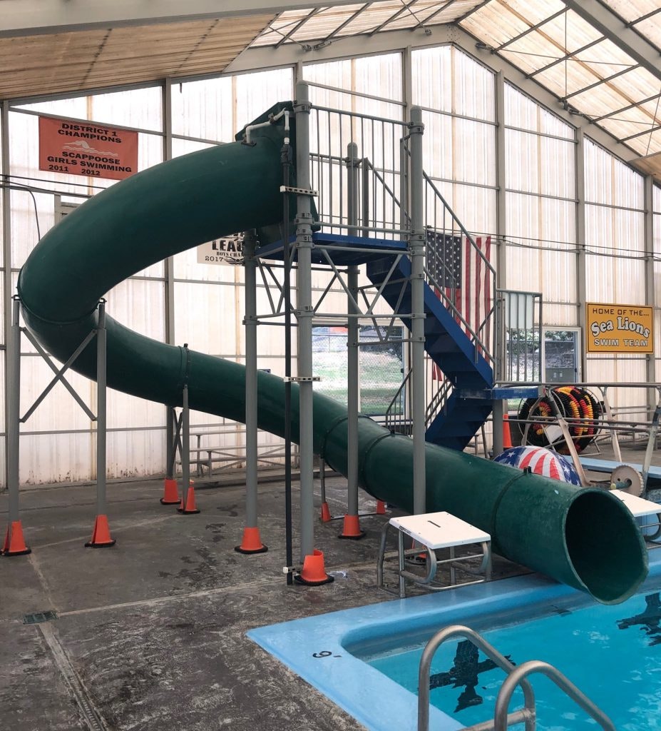 An indoor swimming pool featuring a green water slide, stairs, and diving platforms, alongside various swimming team decorations.