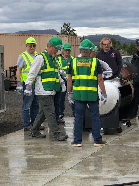 Workers in high-visibility vests and hard hats outdoors near a barrel. (The image is rotated 90 degrees to the left.)