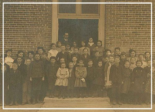 An old sepia photograph of a group of children and teachers posing in front of a brick building, likely a school from an earlier era.