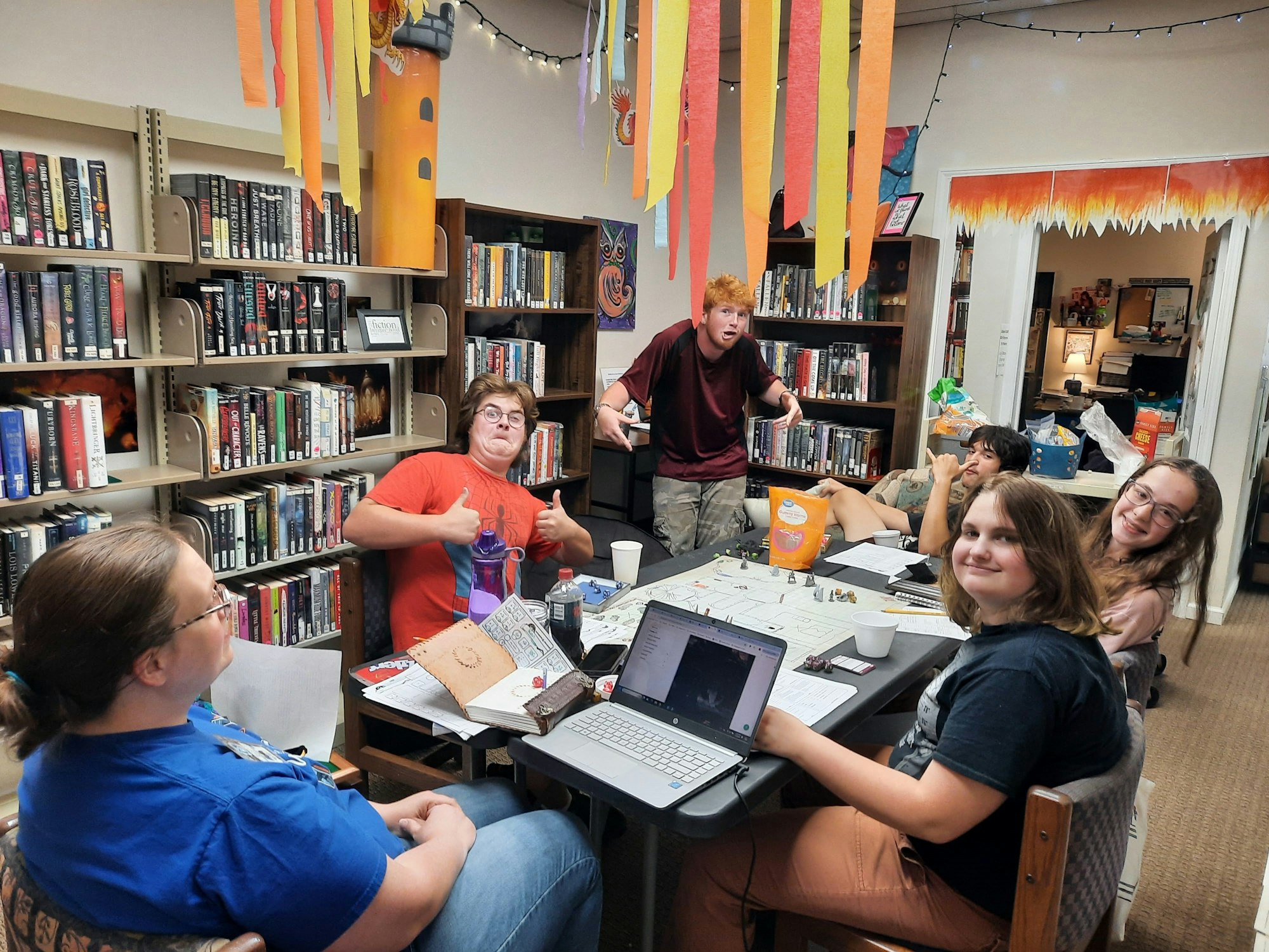 People sitting around a table filled with papers and a laptop, in a room with bookshelves and festive decorations.
