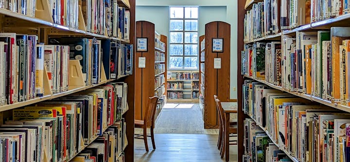 A library aisle with bookshelves full of books and a window at the end.