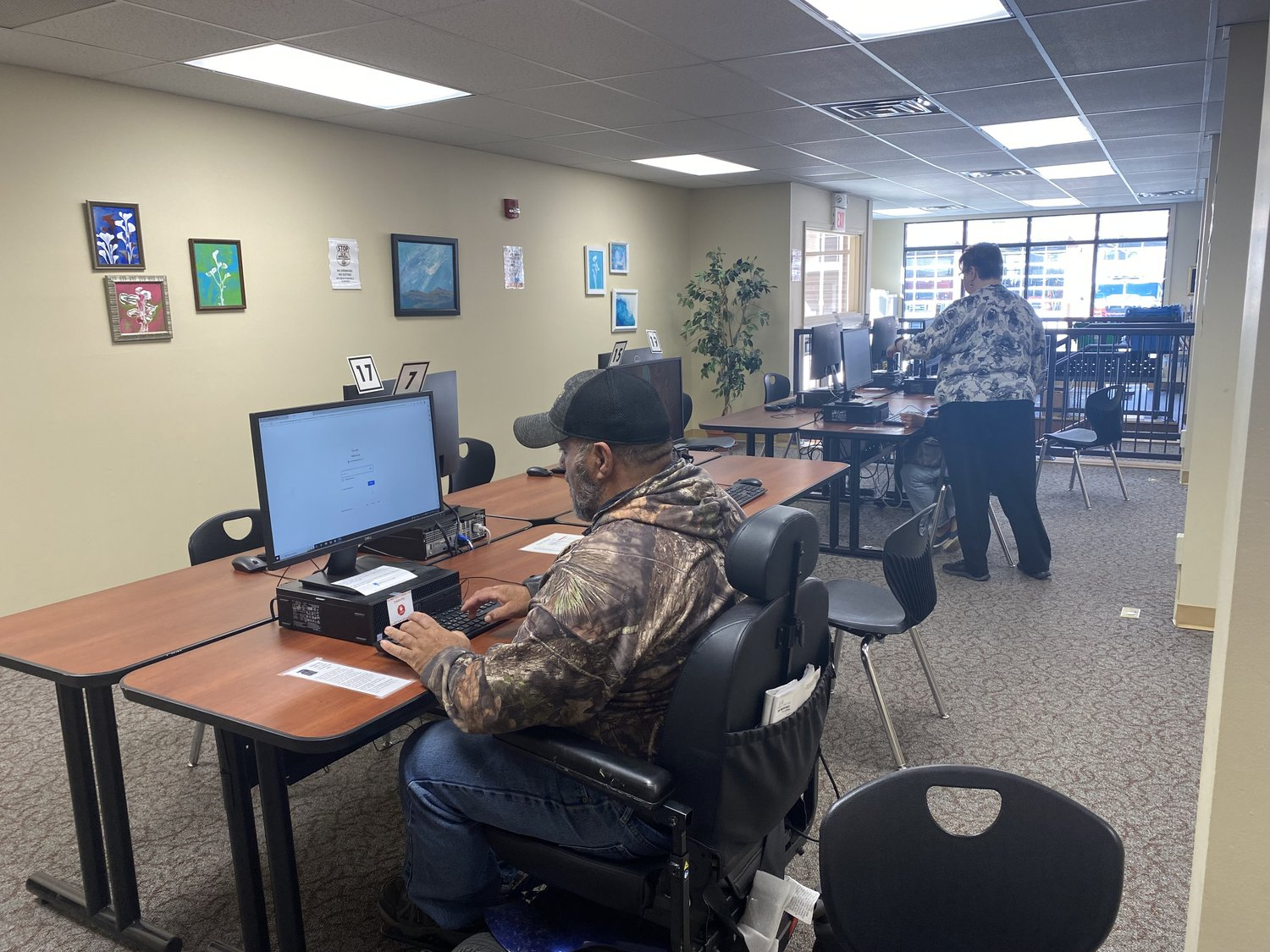 Office with computers and two people, one working and another standing. Art on walls and plants present.
