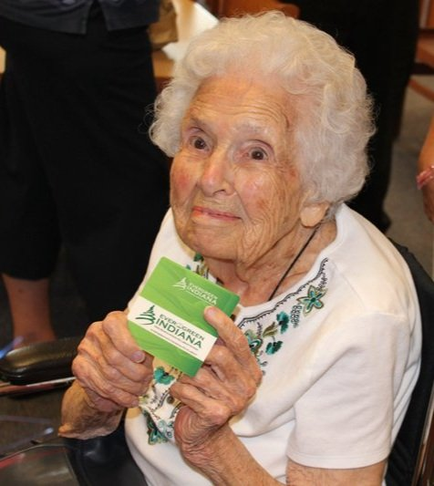 An elderly woman holding a green card or box, smiling, with others partly visible in the background.