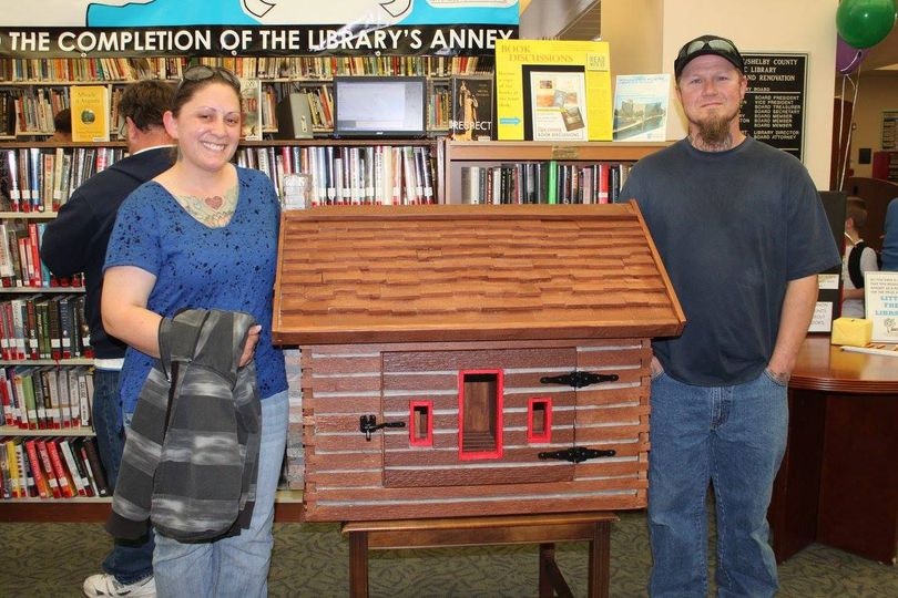 Two people smiling beside a model of a red-door brown cabinetry in a library.