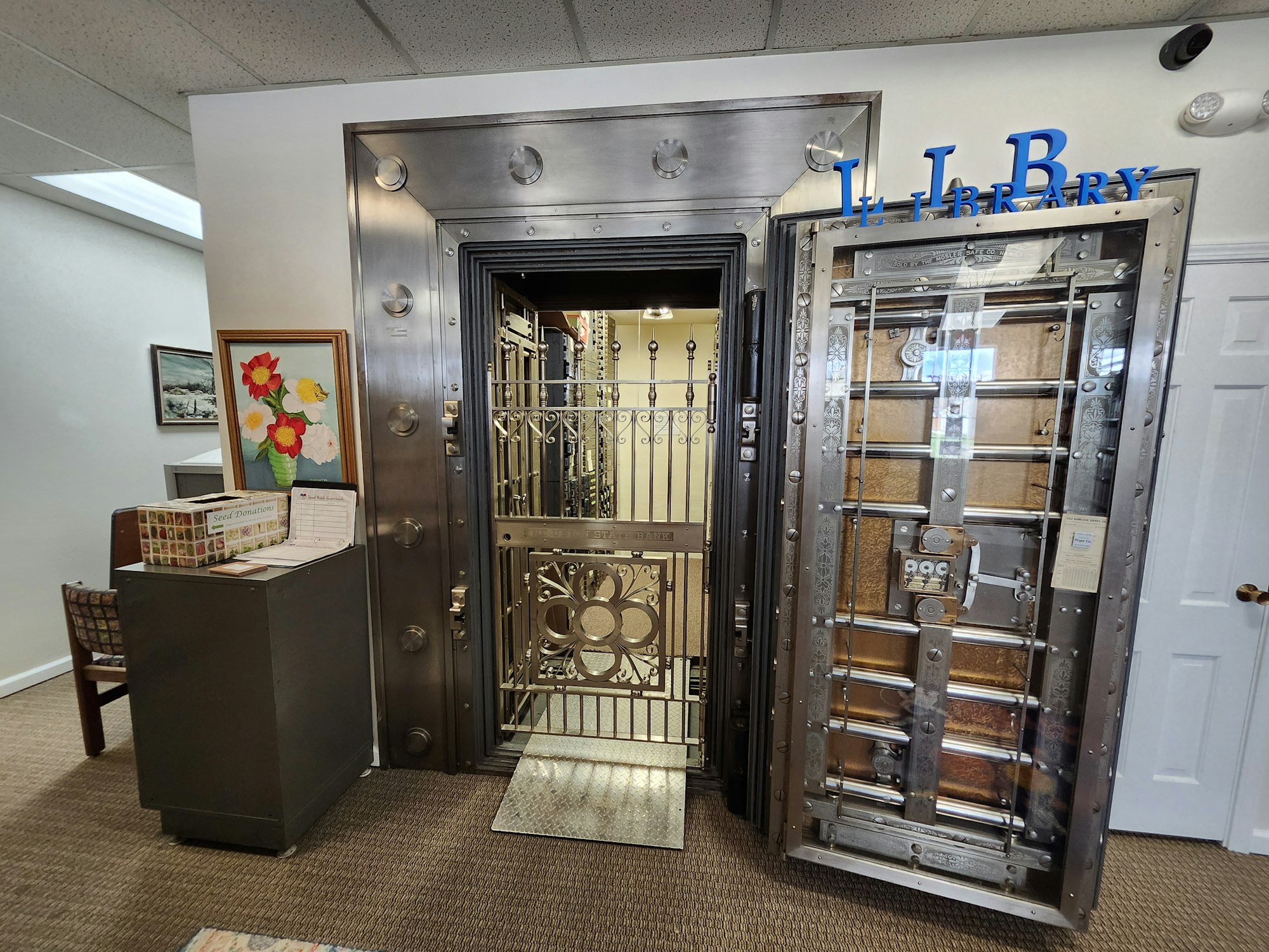 A repurposed vault door opened to a library space, with ornate metalwork, a book display, paintings, and a "LIBRARY" sign.