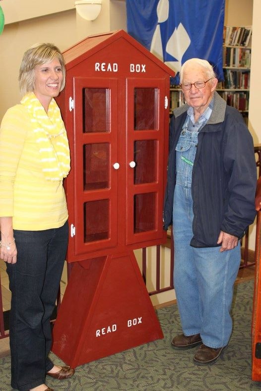 A man and a woman smiling next to a red "READ BOX" book exchange station inside a room.