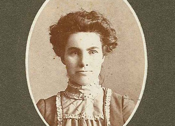 Vintage portrait of a woman with an updo hairstyle, wearing a high-collared ruffled dress, sepia-toned photograph.
