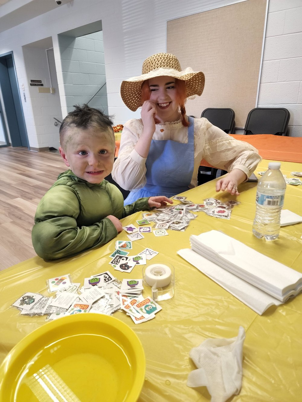 A boy and a woman in a straw hat sitting at a table with stickers, a water bottle, napkins, and a yellow plate.