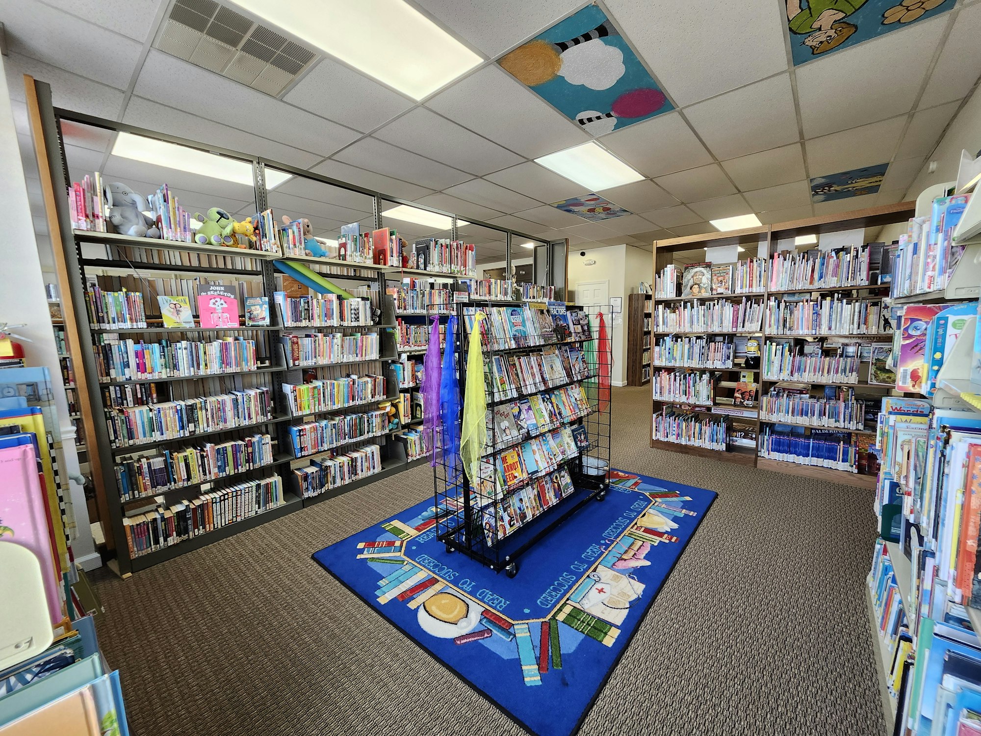 A children's section of a library with books, shelves, and decorative ceiling tiles.