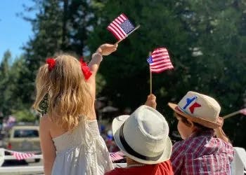 Three children wave American flags, likely at a parade or patriotic event, with one child standing, two seated, in sunny weather.