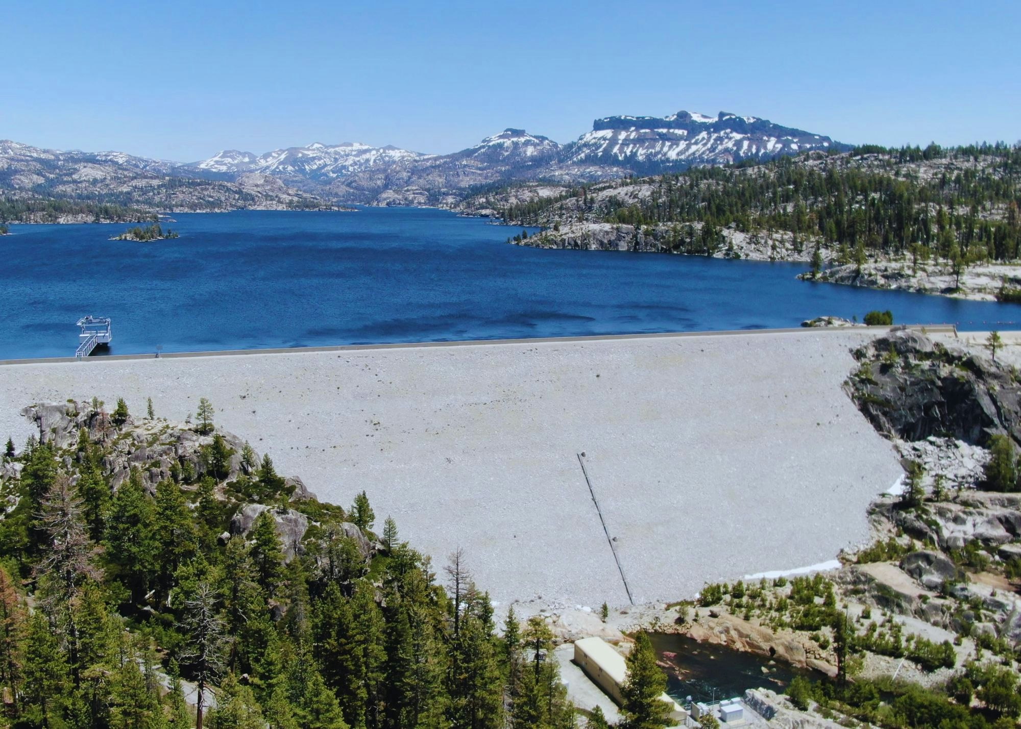 A scenic lake with a dam in front, surrounded by rocky terrain and forested mountains in the background.