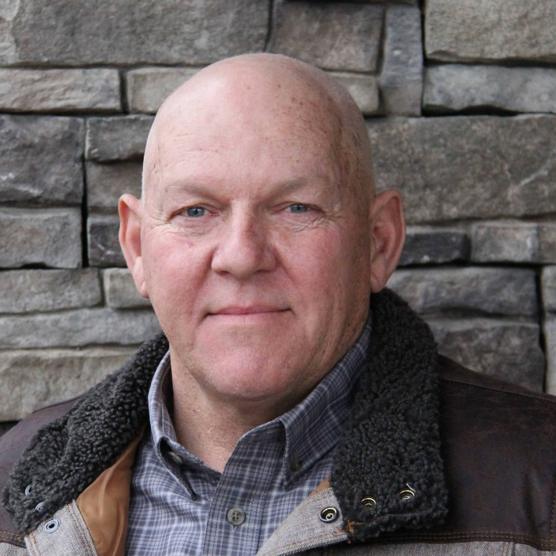 A bald man with blue eyes, wearing a collared shirt and a jacket with a furry collar, in front of a stone wall.