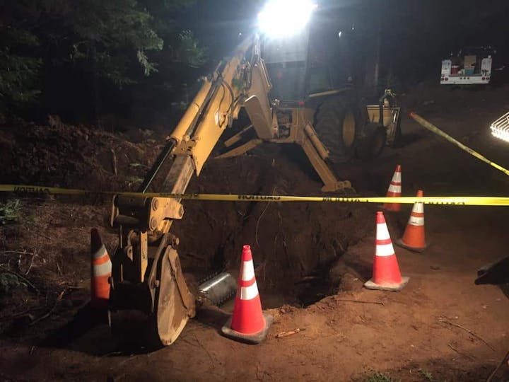 Backhoe at a nighttime construction site with caution tape and traffic cones.