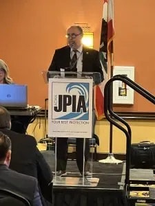 A man is speaking at a podium with a sign reading "JPIA - Your Best Protection.” There are people seated, listening to him.