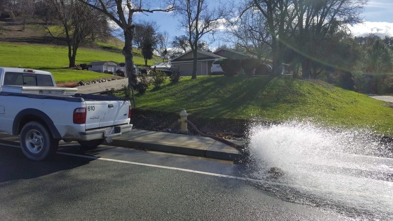 Pickup truck parked beside a fire hydrant spraying water onto a sunny street with greenery around.