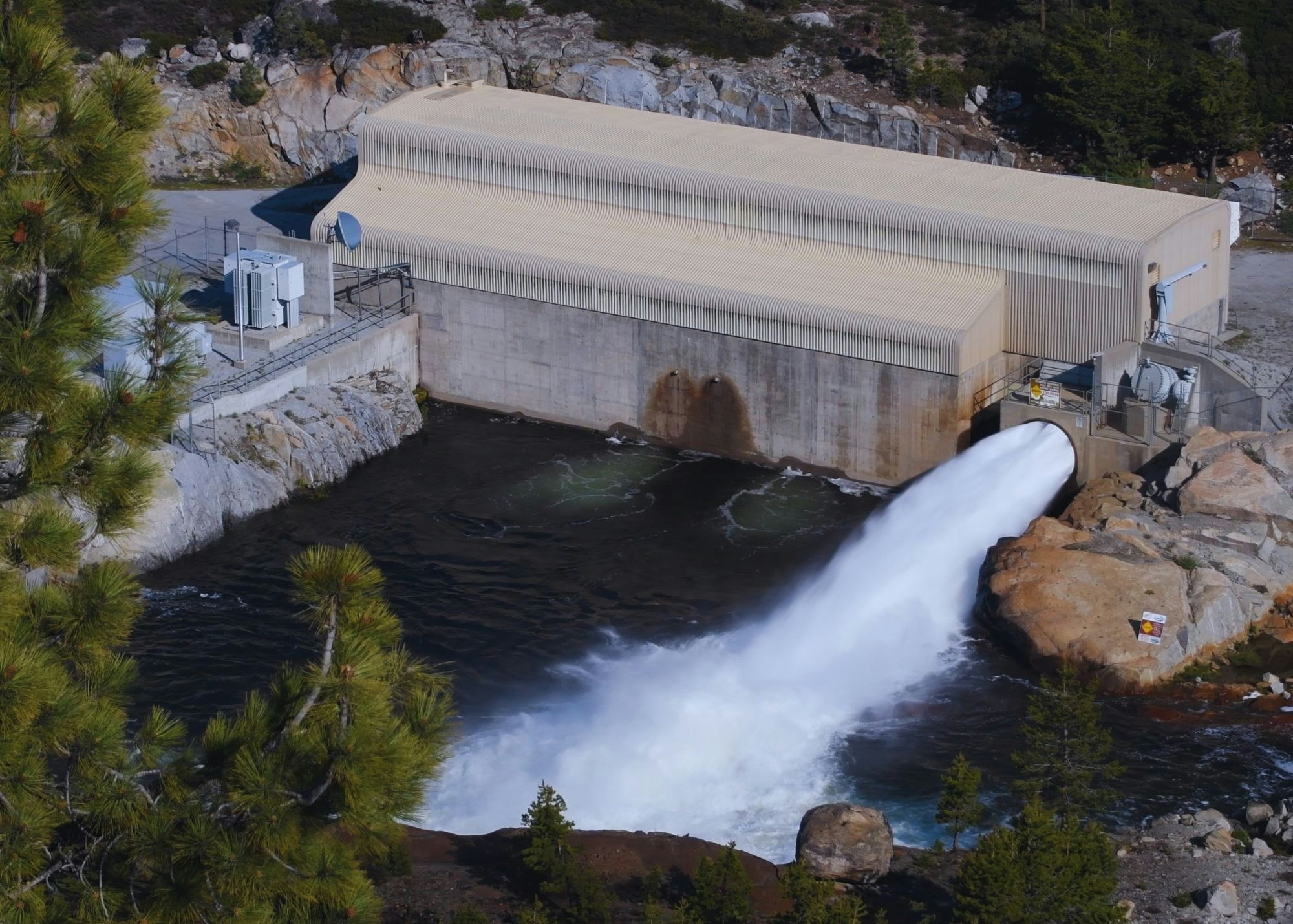 A hydroelectric dam releasing water, surrounded by trees and rocky terrain.