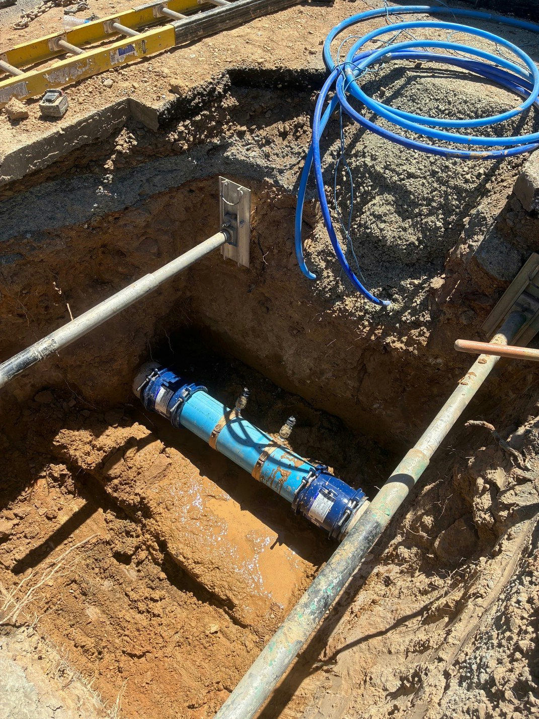 The image shows a construction site with a blue pipe in a trench, surrounded by dirt and tools, including blue hoses and a shovel.