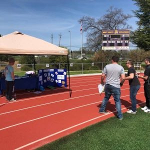 People at a track and field event with a tent, display board, and stadium seats in the background.