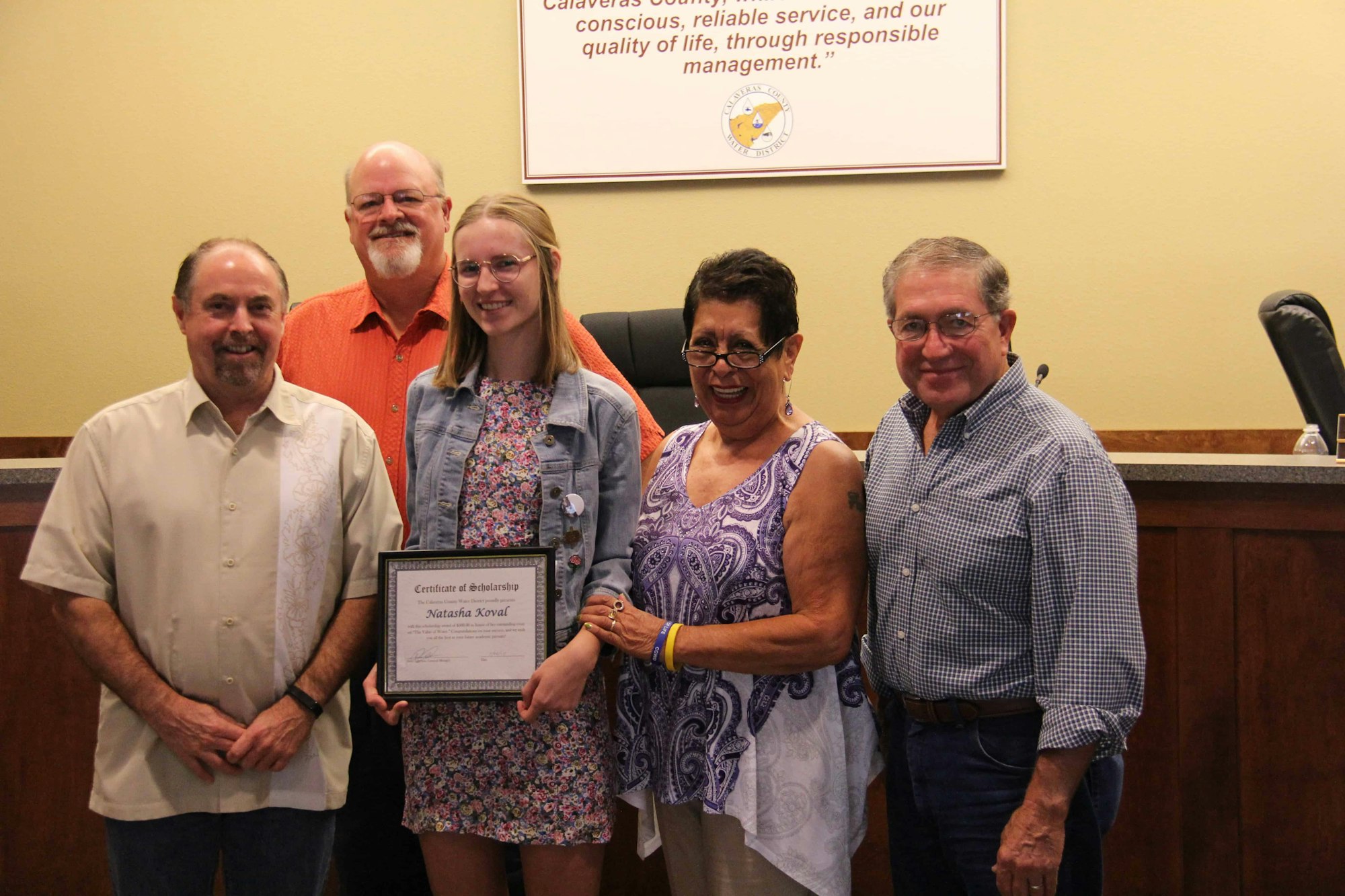 Group of four adults smiling, one holding a "Certificate of Scholarship." Office-like setting with a seal and motto in the background.