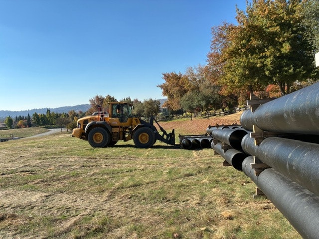 A loader is positioned near large black pipes on grassy land with trees in the background under a clear blue sky.