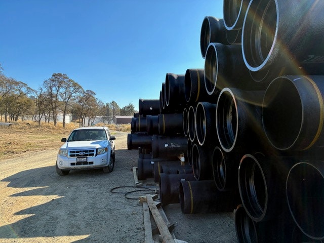 A parked vehicle beside stacks of large black pipes in an outdoor setting with clear skies and trees.