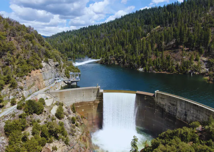 A large dam with a spillway releasing water, surrounded by forested hills and a cloudy sky.
