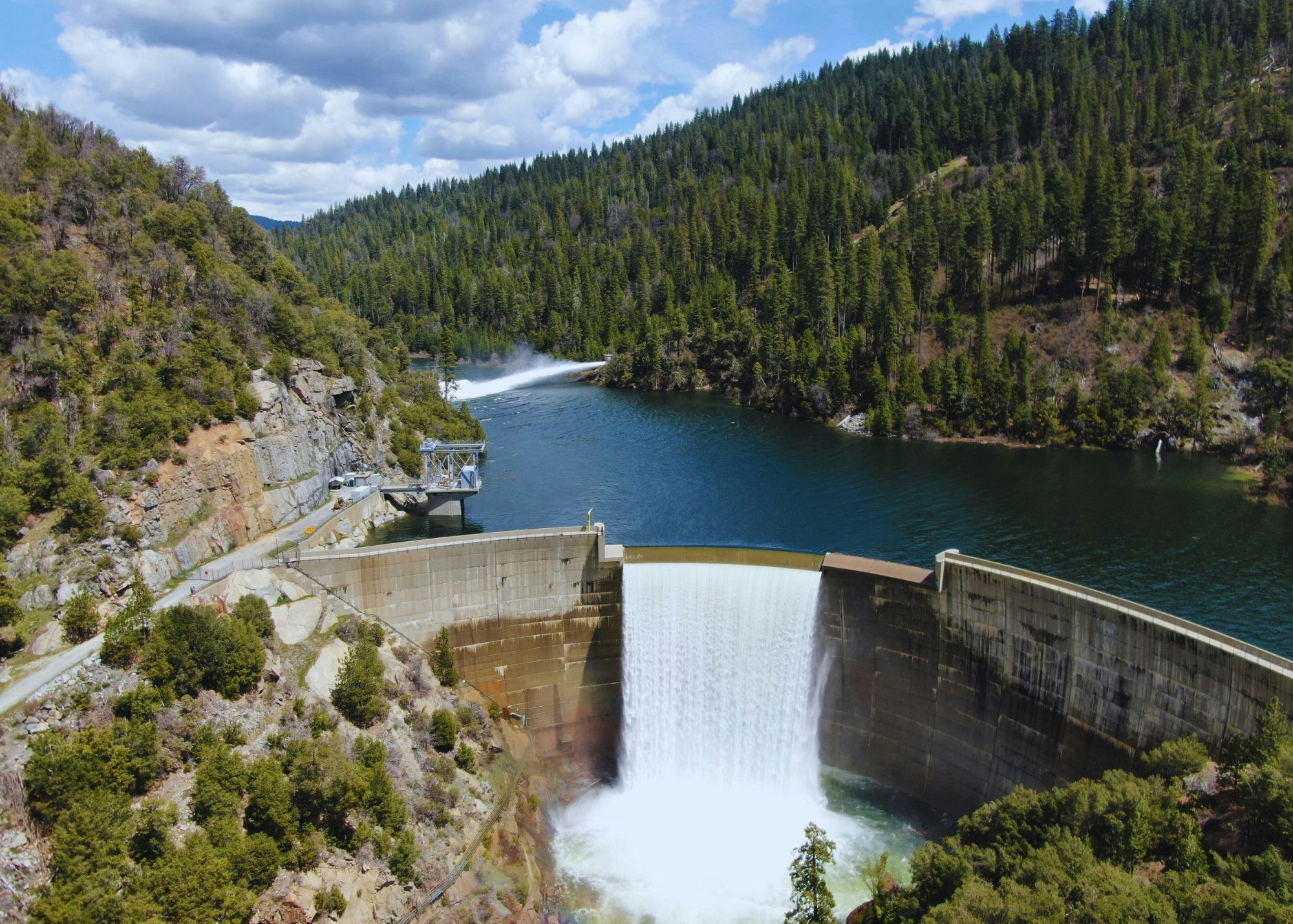 A large dam with a spillway releasing water, surrounded by forested hills and a cloudy sky.